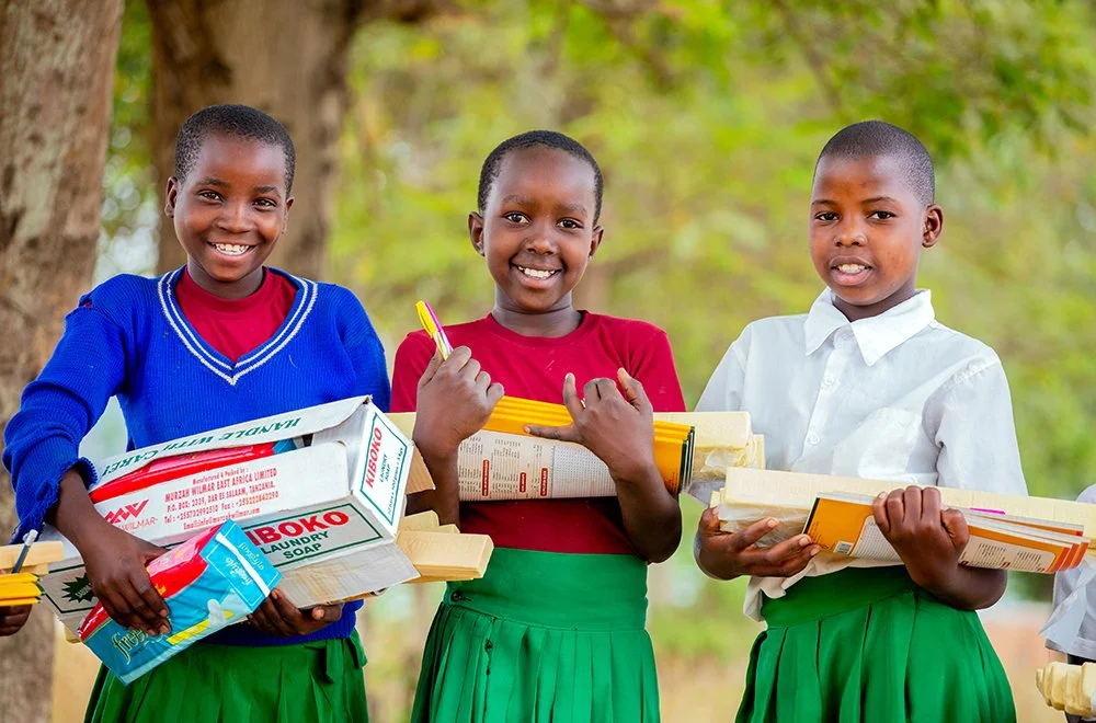 Kolme tansanialaista tyttöä seisoo rinnakkain ja pitelee Planin jakamia hygieniatarvikkeita.