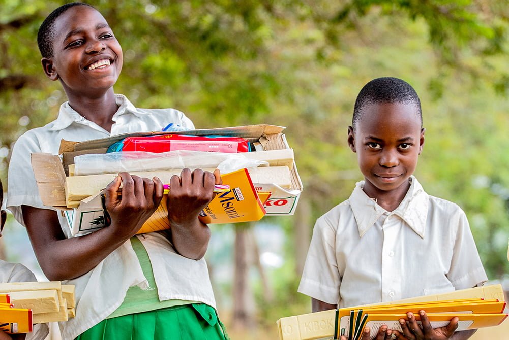 Kaksi tansanialaista tyttöä hymyilee ja kantaa sylissään tarvikkeita.