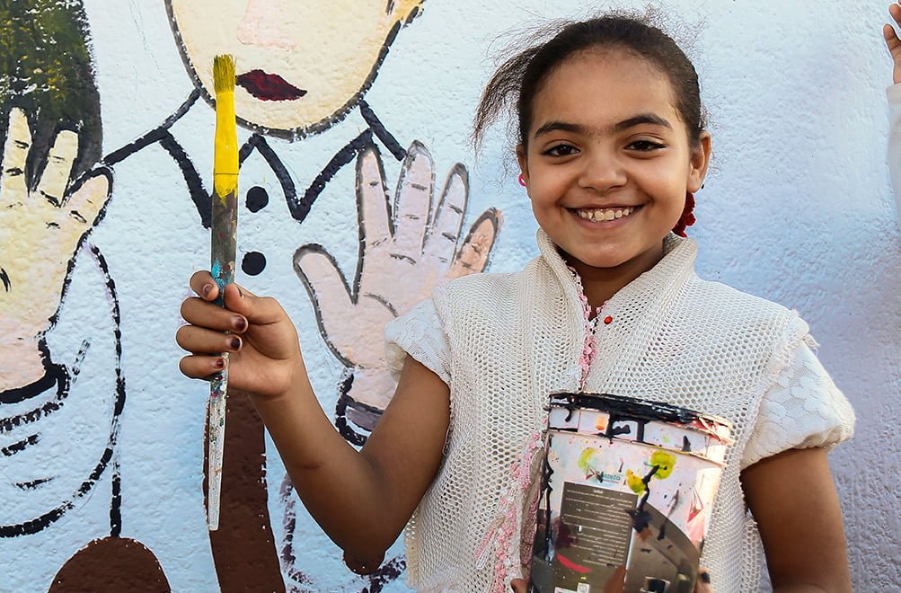 Tyttö seisoo pensseli ja maalipurkki kädessään värikkään seinän edessä.