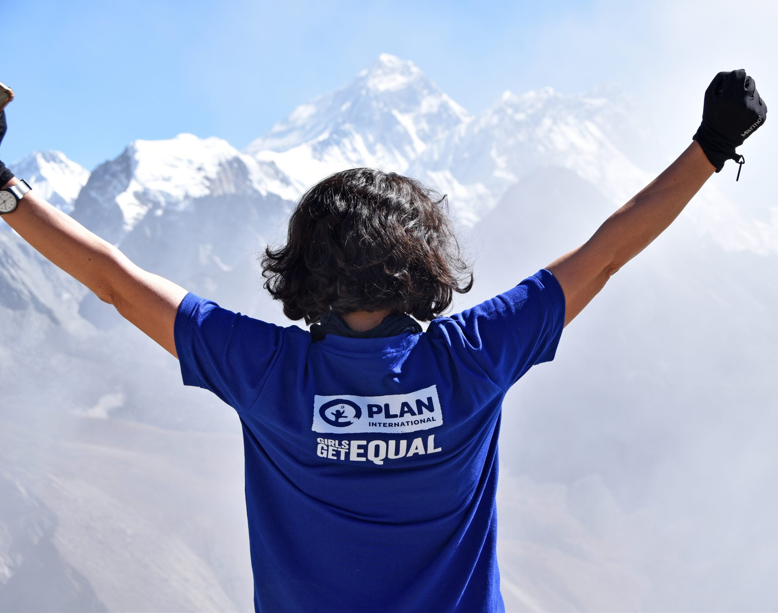 Takaapäin kuvattu henkilö katsoo vuoristomaisemaa ja pitää käsiään ylhäällä kuin voiton merkiksi.