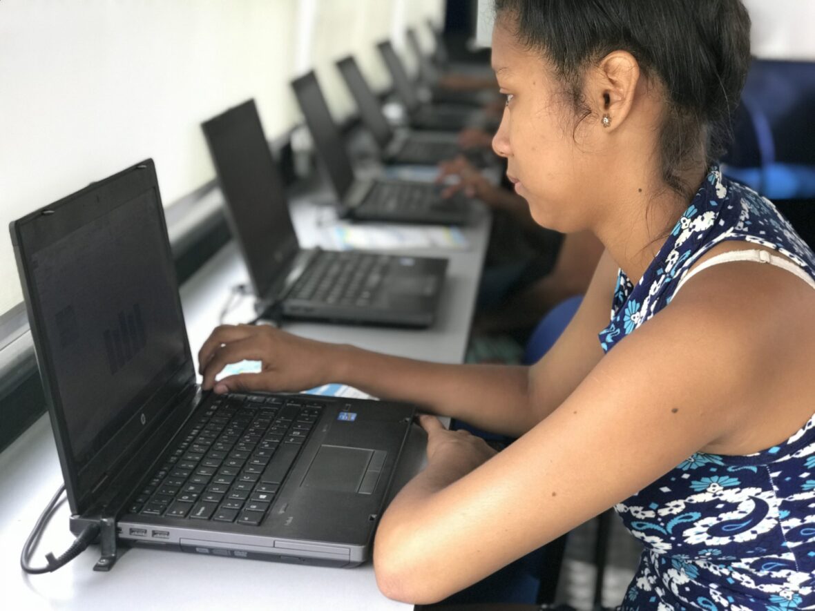 Tyttö käyttää tietokonetta, taustalla pitkä rivi tietokoneita.