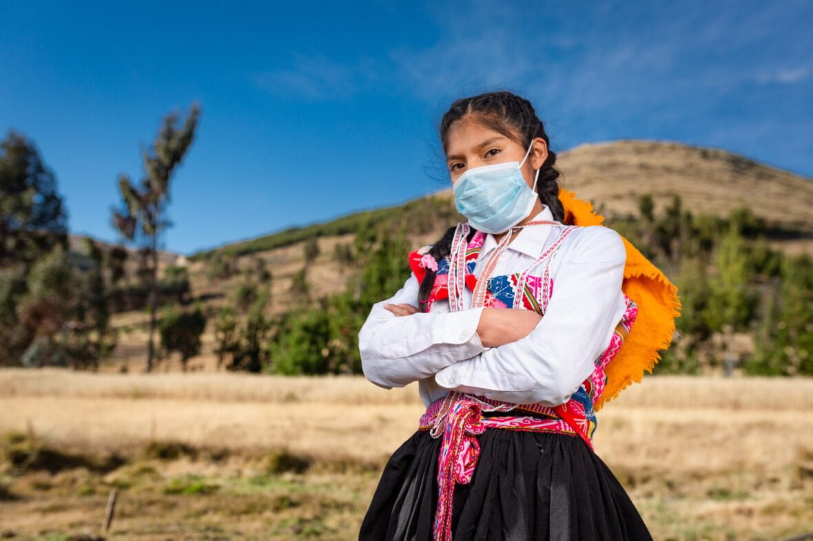 Perulainen tyttö seisoo kädet puuskassa, maski kasvoilla ja koulureppu selässä.