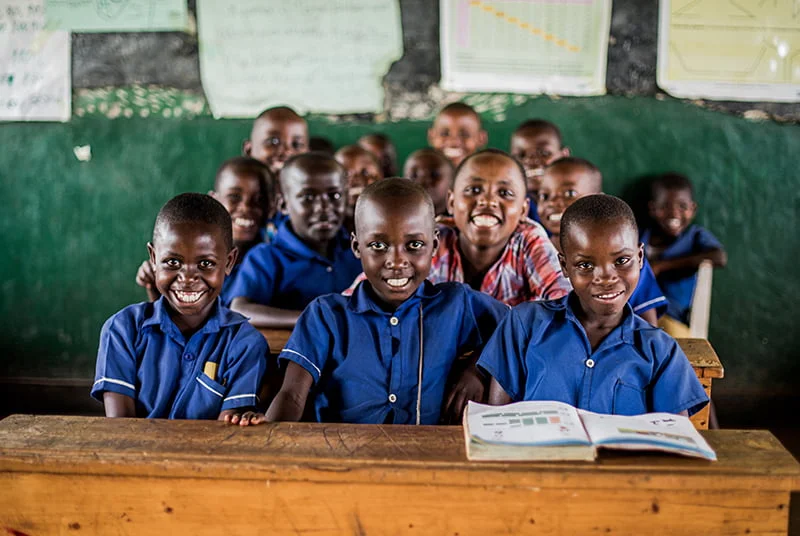 Ryhmä ruandalaisia lapsia istuu koululuokassa sinisissä koulupuvuissa ja katsoo hymyillen kohti kameraa.
