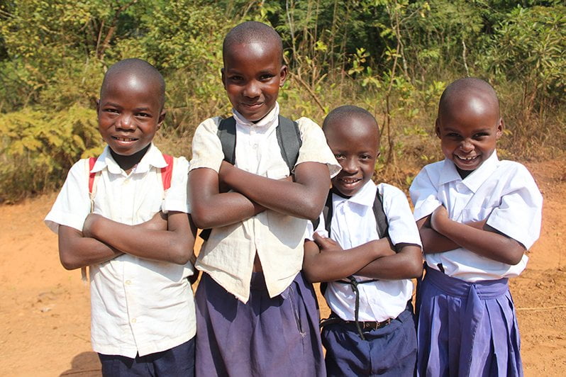 Tansanialaiset lapset seisovat ulkona koulupuvuissa kädet puuskassa ja katsovat kohti kameraa.