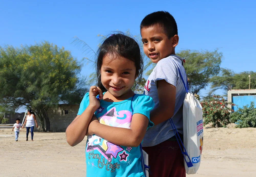 Aukiolla seisoo kaksi perulaista lasta, joista toisella on selässään Planin antama koulutarvikepakkaus.
