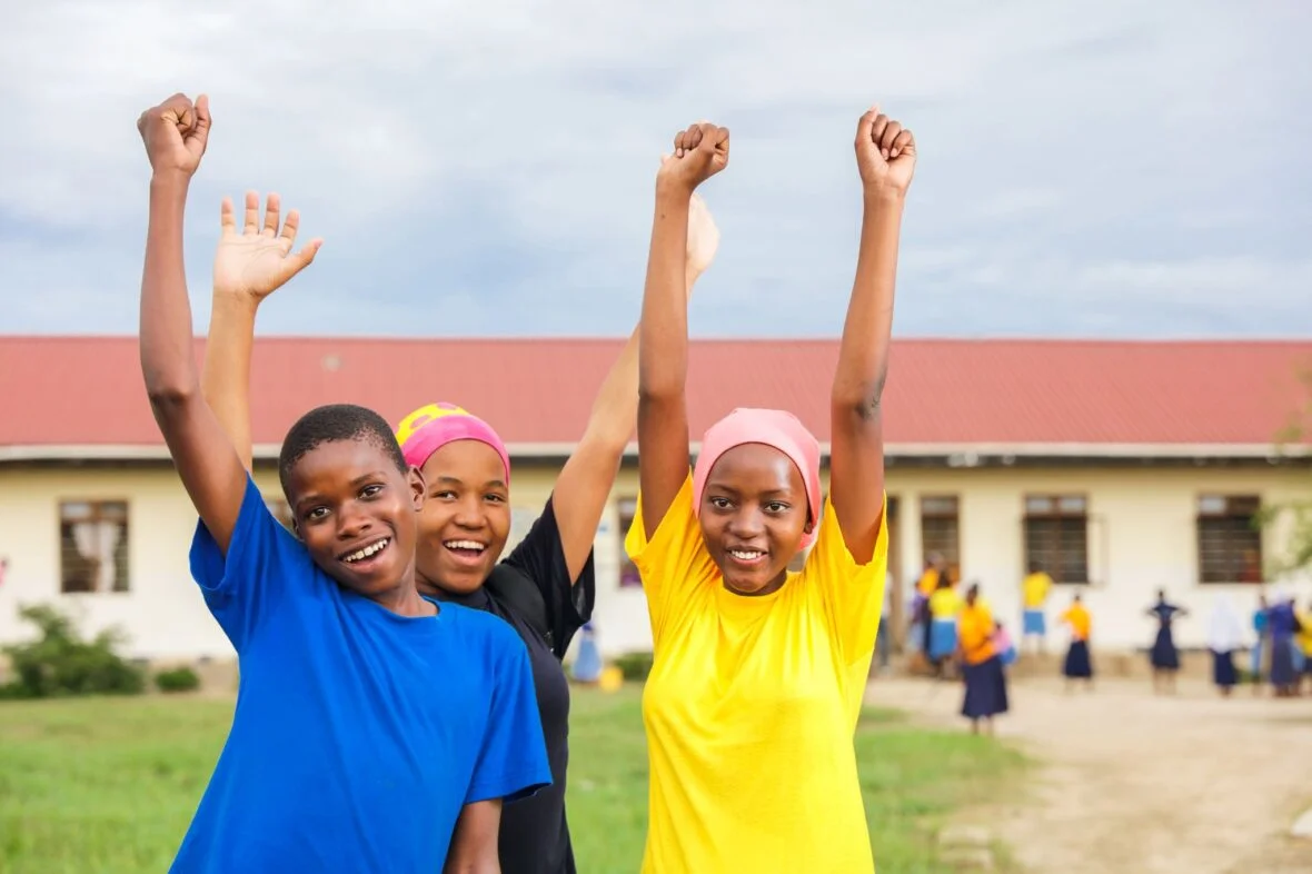 Kolme tansanialaistyttö iloisina kädet ylhäällä koulun pihalla.
