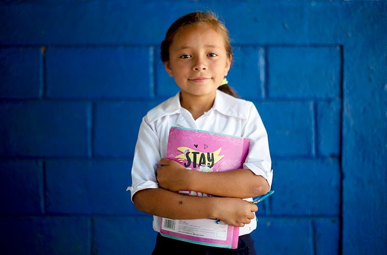 Nicaragualainen tyttö seisoo sinisen seinän edessä kirja sylissään.