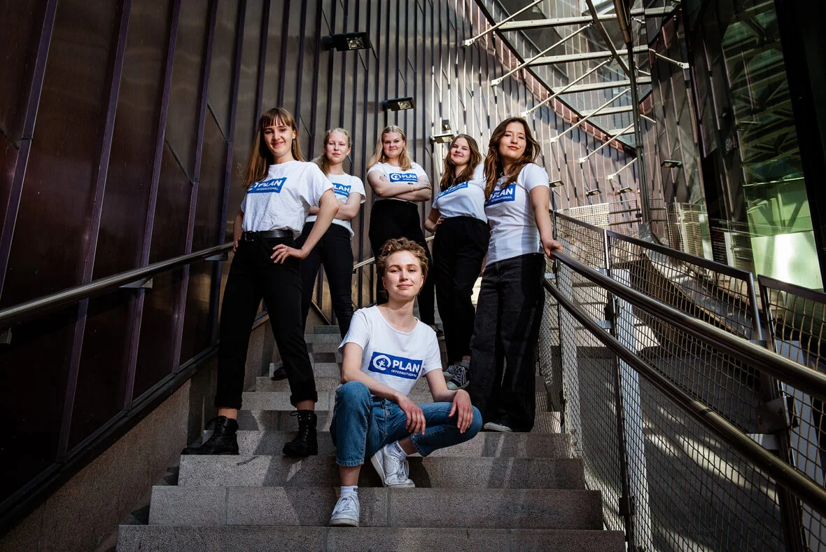 Kuusi nuorta tyttöä Planin t-paidoissa seisoo rappusissa ja poseeraa kameralle.