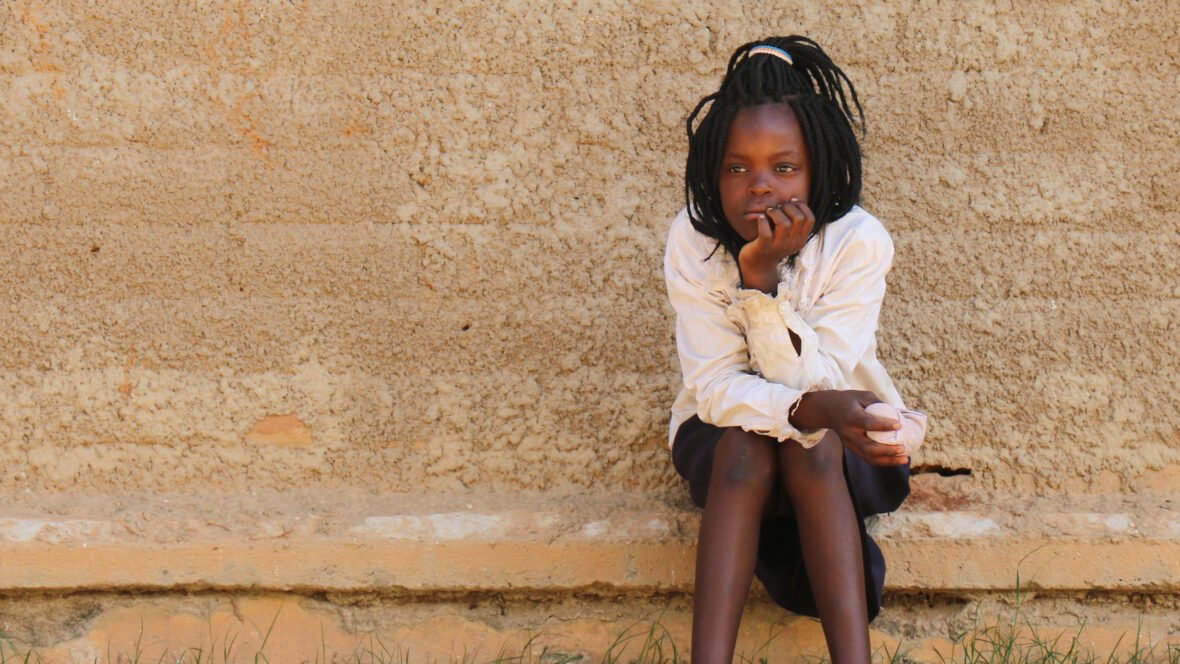 Sambialainen tyttö istuu mietteliäänä penkillä tiiliseinän edustalla.