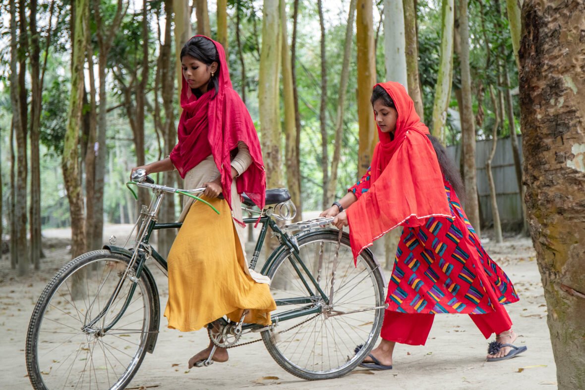 Bangladeshilainen tyttö pyöräilee ja toinen tyttö kävelee pyörän perässä.
