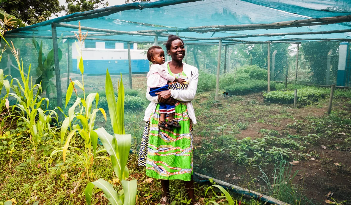 Ugandalainen Evelyne lapsensa kanssa kasvihuoneen edessä.