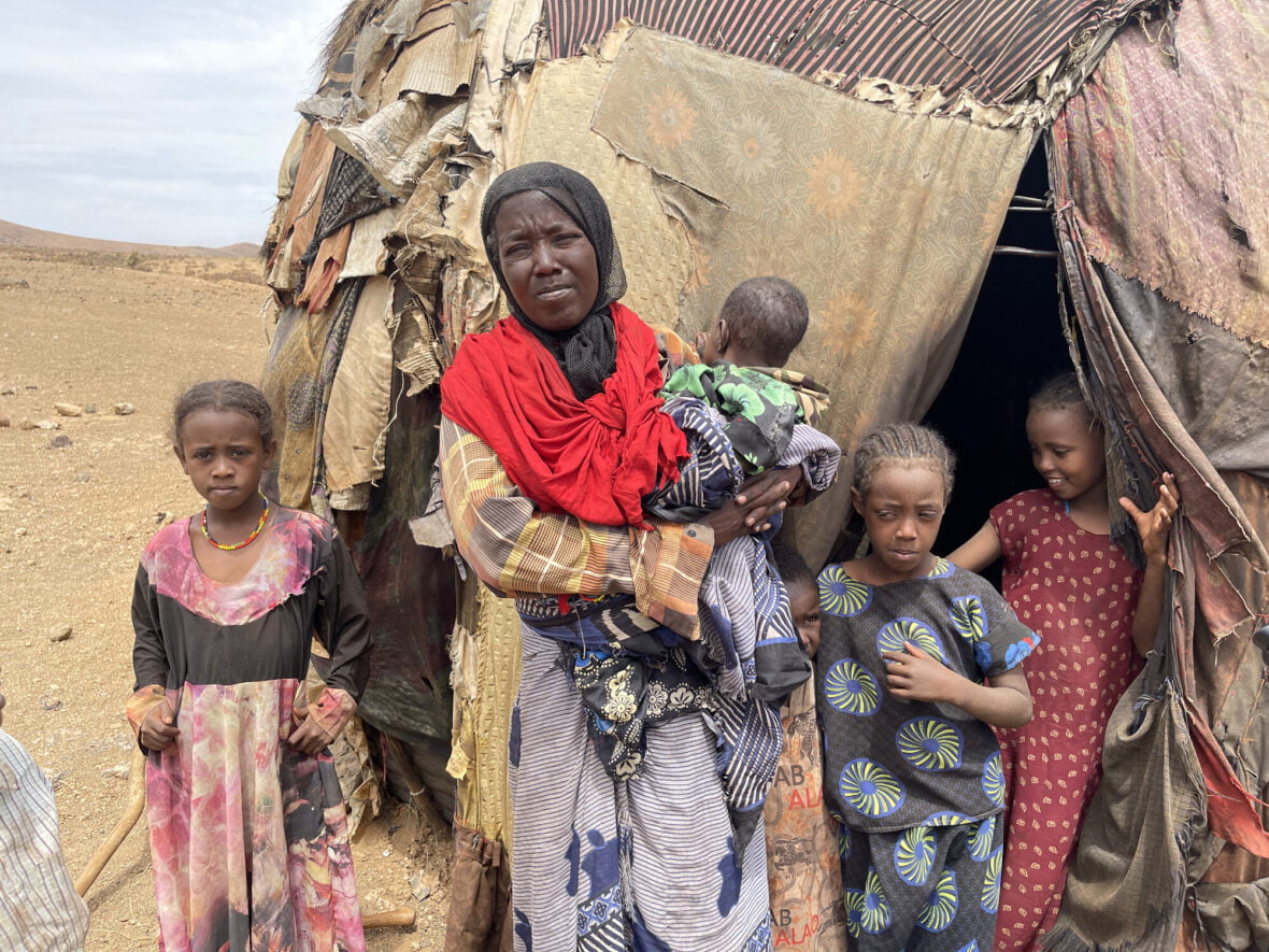 Nainen seisoo kotikutoisen teltan edessä keskellä aavikkoa. Sylissään hänellä on pieni vauvaa ja vierellään useita pienempiä lapsia. 