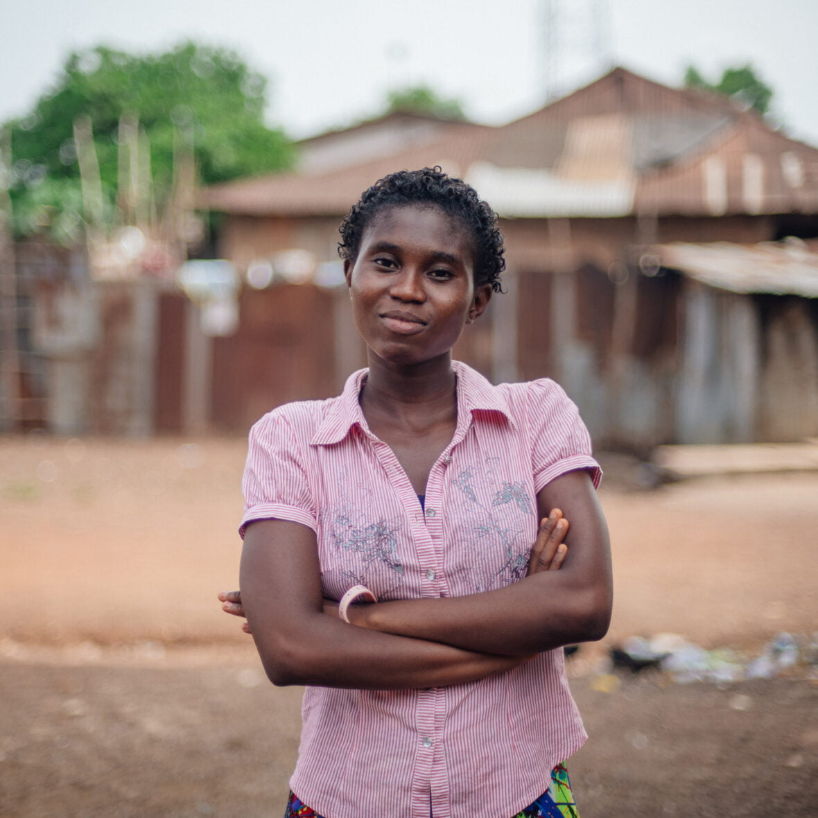 Sierraleonelainen tyttö seisoo kotikylänsä edessä ja katsoo kameraan, hän on ristinyt kädet eteensä.