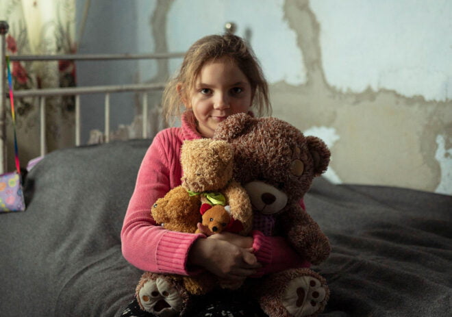 Ukrainalainen tyttö istuu sängyllä halaten nalleja.