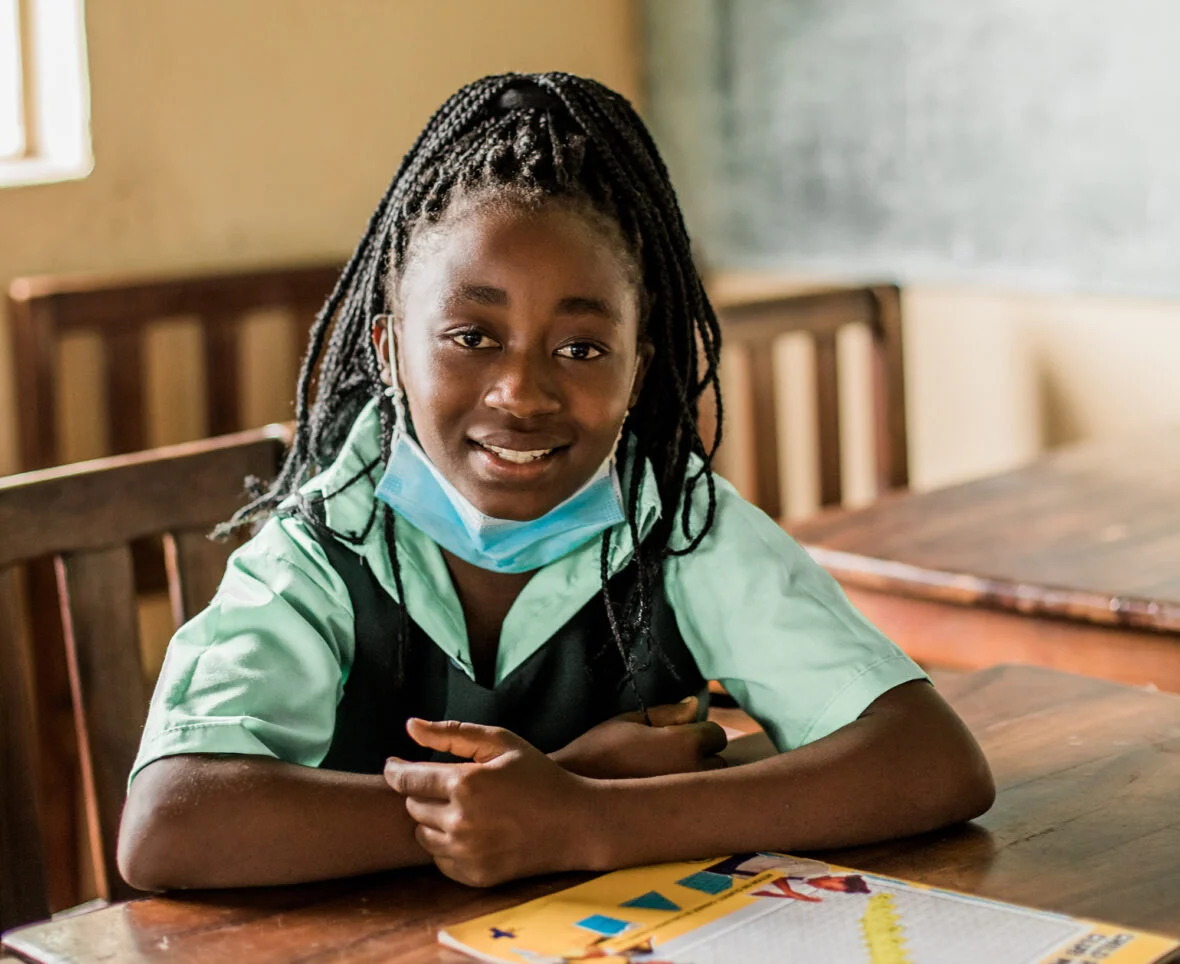 Sambialainen tyttöä istuu koululuokassa. Tytöllä on edessään koulukirja ja hän katsoo suoraan kameraan ja hymyilee. Plan International tukee tyttöjen koulunkäyntiä, myös kuukautisten aikana. On tärkeää, että tytöt voivat käydä koulussa myös silloin, kun heillä on kuukautiset.