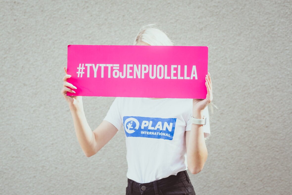 Henkilö Planin t-paidassa pitää edessään pinkkiä kylttiä, jossa lukee #tyttöjenpuolella.