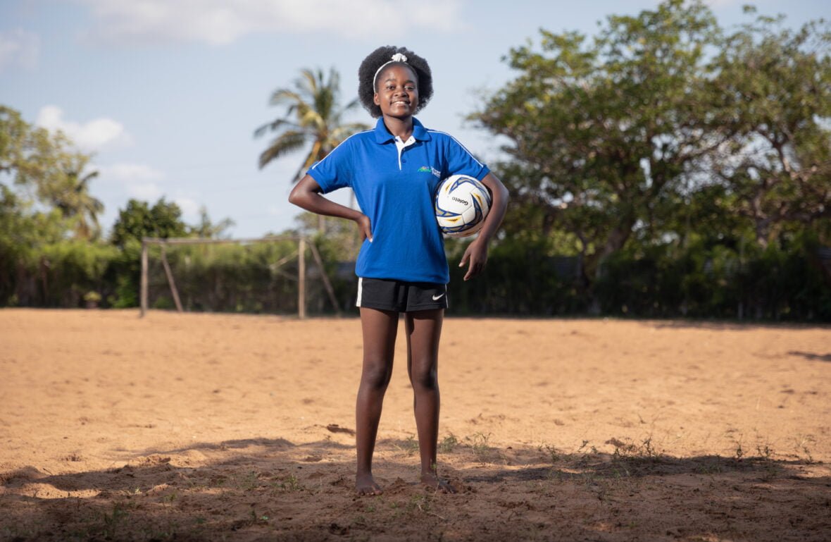 Mosambikilainen Eunice poseeraa jalkapallokentällä, toinen jalka jalkapallon päällä. Eunice katsoo suoraan kameraan päättäväisesti. Hän on ristinyt kädet eteensä. Hänen takanaan näkyy puita, kuten palmuja.
