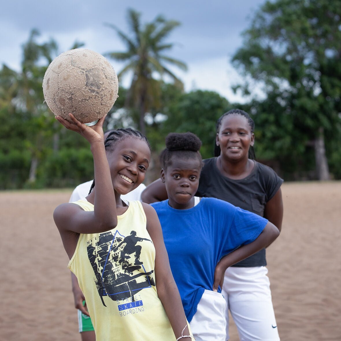 Kolme mosambikilaista tyttöä poseeraa keskellä hiekkaista jalkapallokenttää. Lähempänä kameraa olevalla tytöllä on yllään keltainen t-paita ja kädessään, kohotettuna päänsä ylepäpuolelle, jalkapallo. Kaikki tytöt katsovat suoraan kameraan. Jalkapalloa pitelevä tyttö hymyilee leveästi kameralle.