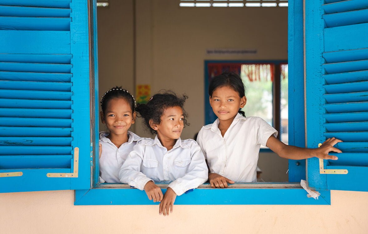 Kolme kambodzalaista tyttöä kurkkaa ulos avoimesta koulurakennuksen ikkunasta. Tytöillä on yllään valkoiset koulupuvun paidat. Kaksi kolmesta tytöstä katsoo suoraan kameraan, ja hymyilee. Keskimmäinen tytöistä katsoo vasemmalle hymyillen. Ikkunassa on kirkkaan turkoosit karmit.