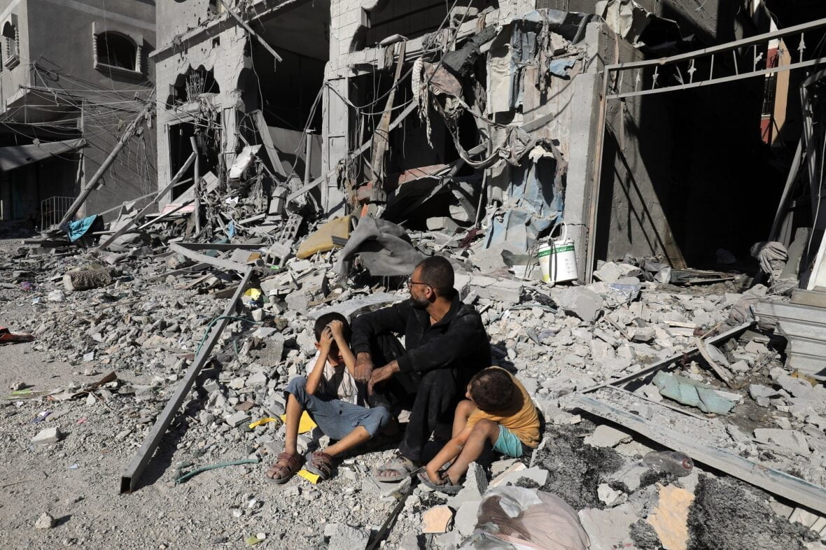 Mies ja kaksi lastaa istuu täysin tuhoutuneiden raunioiden keskellä.