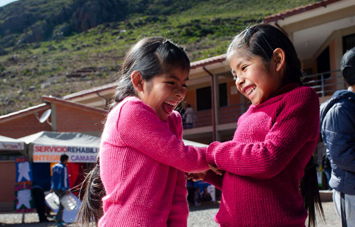 Valokuvassa on kaksi pinkkeihin neulepaitoihin pukeutunutta pientä tyttöä. Tytöt ovat kääntyneet hieman kamerasta pois päin. He pitävät toisiaan käsistä ja nauravat iloisesti, silmät suljettuina. Tyttöjen taustalla näkyy kylää ja kylän taustalla vuoristomaista maisemaa.