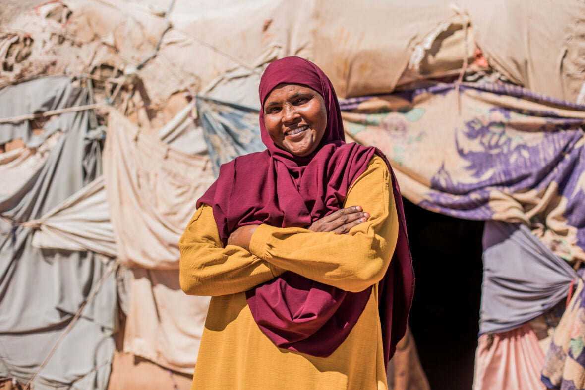 Nainen seisoo kankaista ja puusta rakennetun kotinsa edessä kädet puuskassa ja itsevarmasti hymyillen pakolaisleirillä Somaliassa.