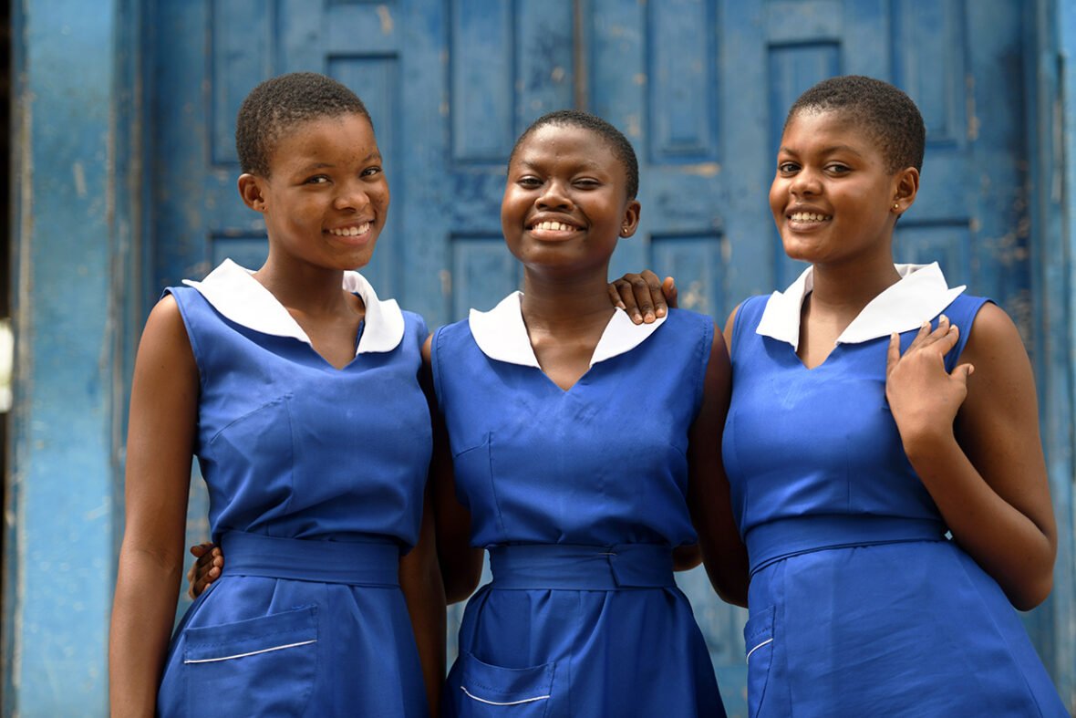 Kolme ghanalaista tyttöä seisoo rinnakkain, kädet toistensa hartioille. He katsovat suoraan kameraan ja hymyilevät iloisesti.