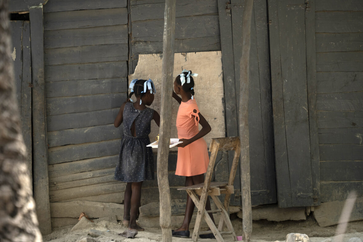 Kaksi tyttöä Haitissa seisovat rakennuksen edessä ja kirjoittavat rakennuksen seinässä olevaan puutauluun.