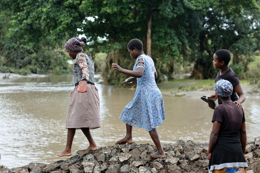 Kolme tyttöä ylittää kävellen tulvan tuhoamaa kivisiltaa.