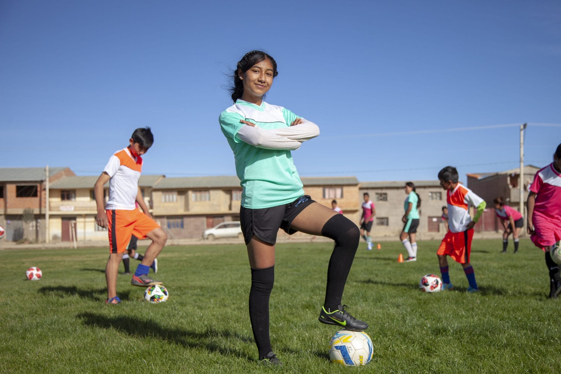 Nuori tyttö poseeraa jalkapallokentällä jalkapallon kanssa itsevarmasti kädet puuskassa.