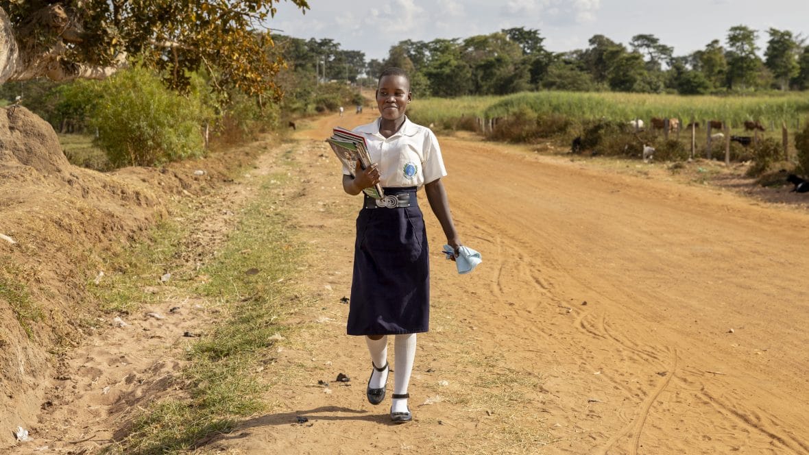 Ugandalainen tyttö kävelee hiekkatietä pitkin kirjat kädessään.