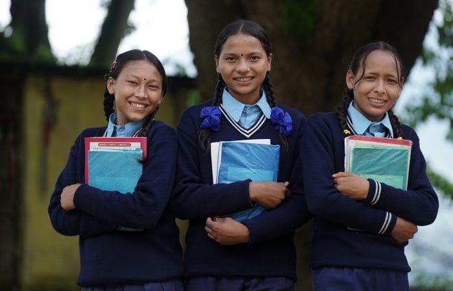 Kolme koulutyttöä seisoo ja katsoo kameraan, heillä on sylissään koulukirjoja.