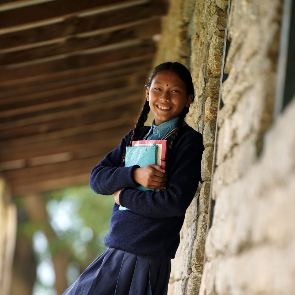 Nepalilainen tyttö nojaa koulun seinään, sylissään hänellä on koulukirjoja.