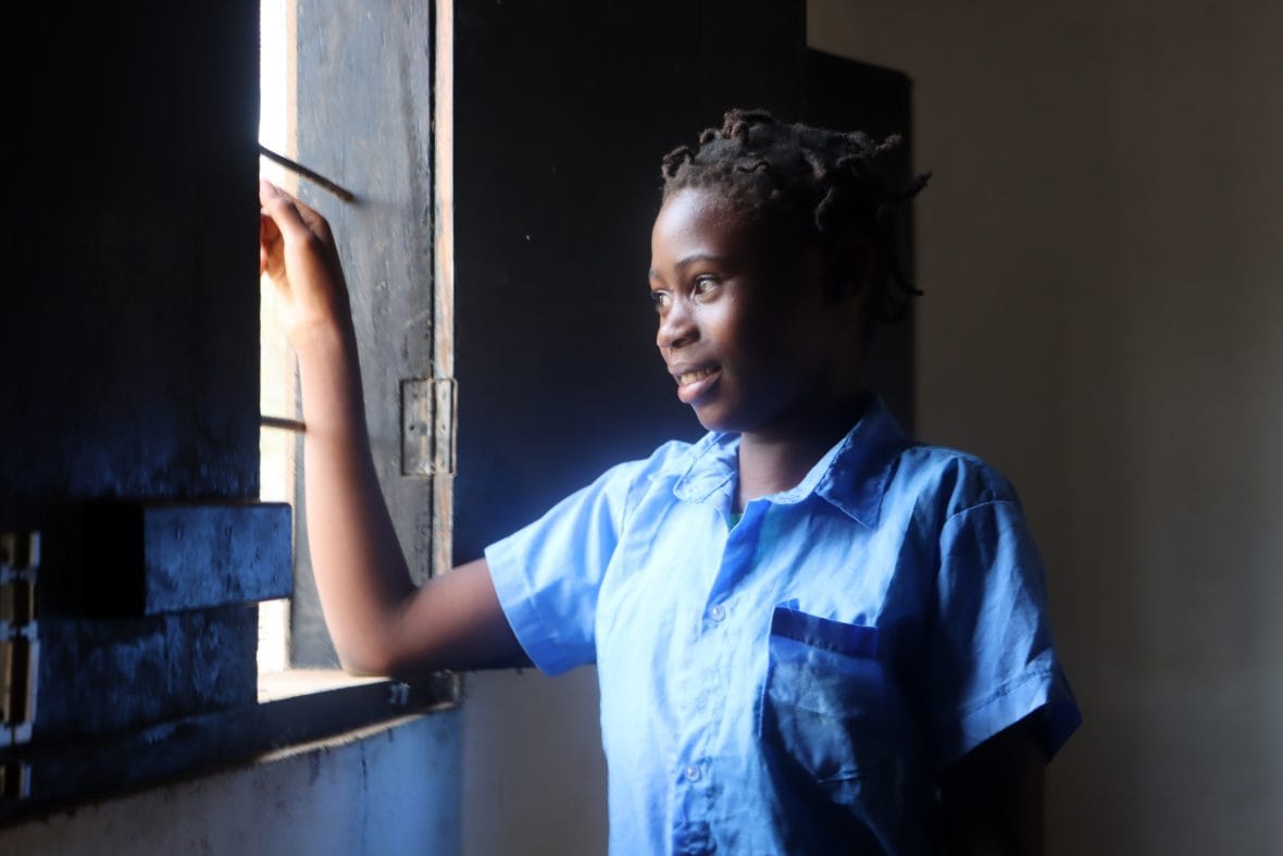 Nuori tyttö seisoo hämärässä huoneessa ja katsoo valoisasta ikkunasta ulos.