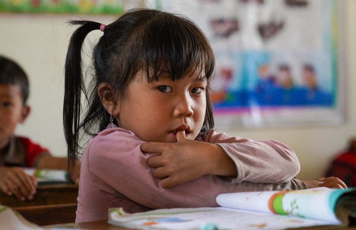 Nuori tyttö koululuokassa katsoo kameraan ja nojaa pulpettiin edessään.