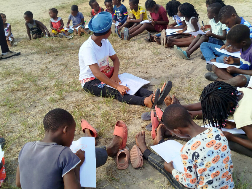 Joukko lapsia istuu ringissä paljaalla maalla.