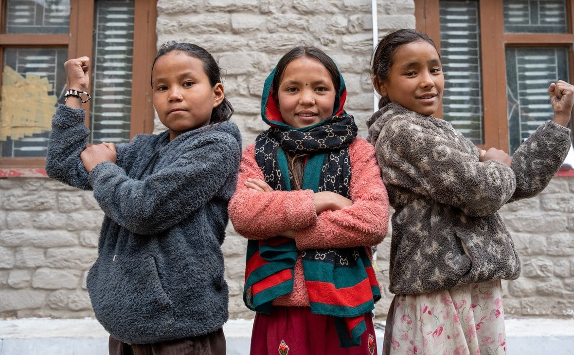 Kolme nepalilaista tyttöä seisoo voimakkaissa asennoissa vierekkäin ja katsoo kameraan.